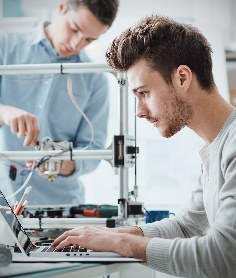Un estudiante ajustando una impresora 3D mientras otro teclea en un ordenador portátil